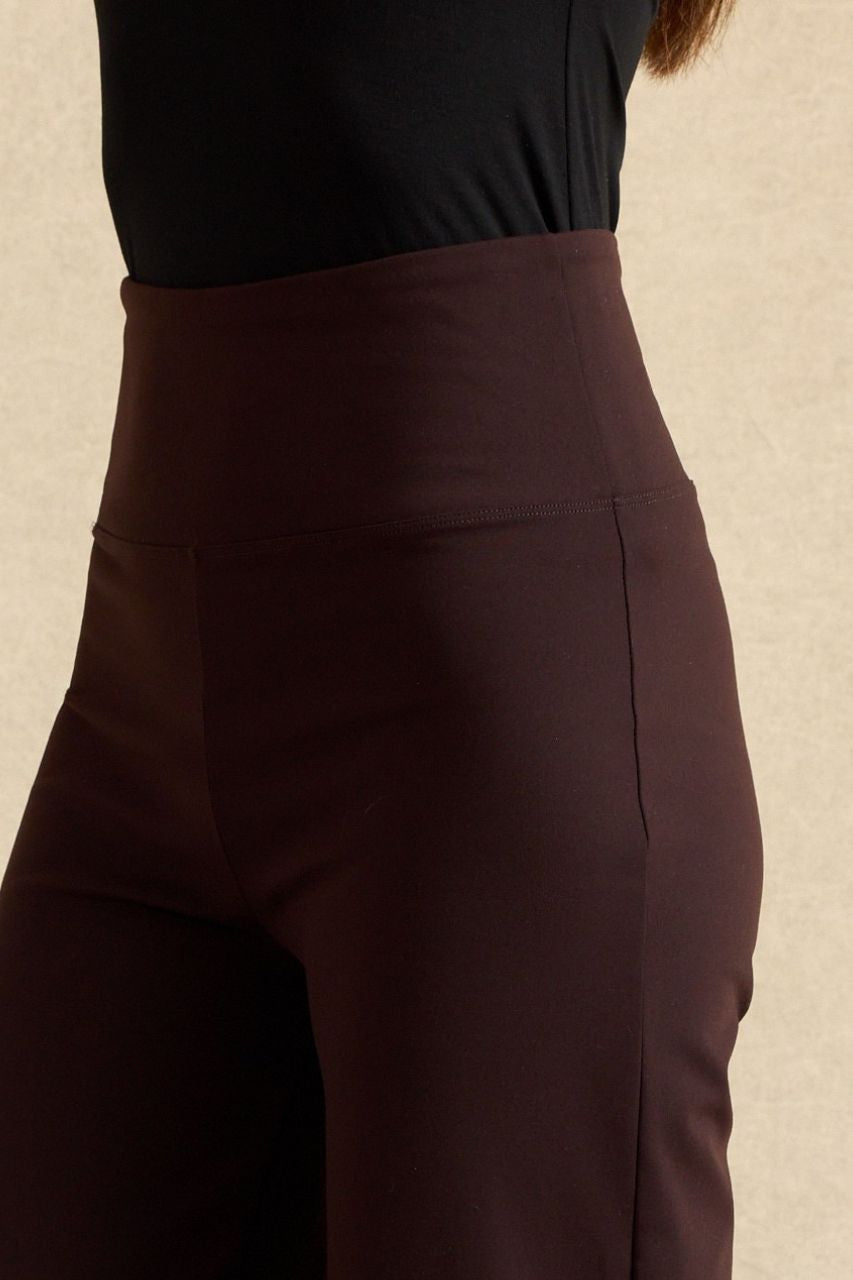 Close-up of a person wearing high-waisted dark brown leggings or pants with a black top, against a light beige textured background.