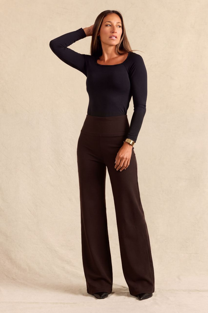 A woman modeling a black long-sleeve scoop-neck top and dark brown wide-leg pants against a textured beige studio backdrop.