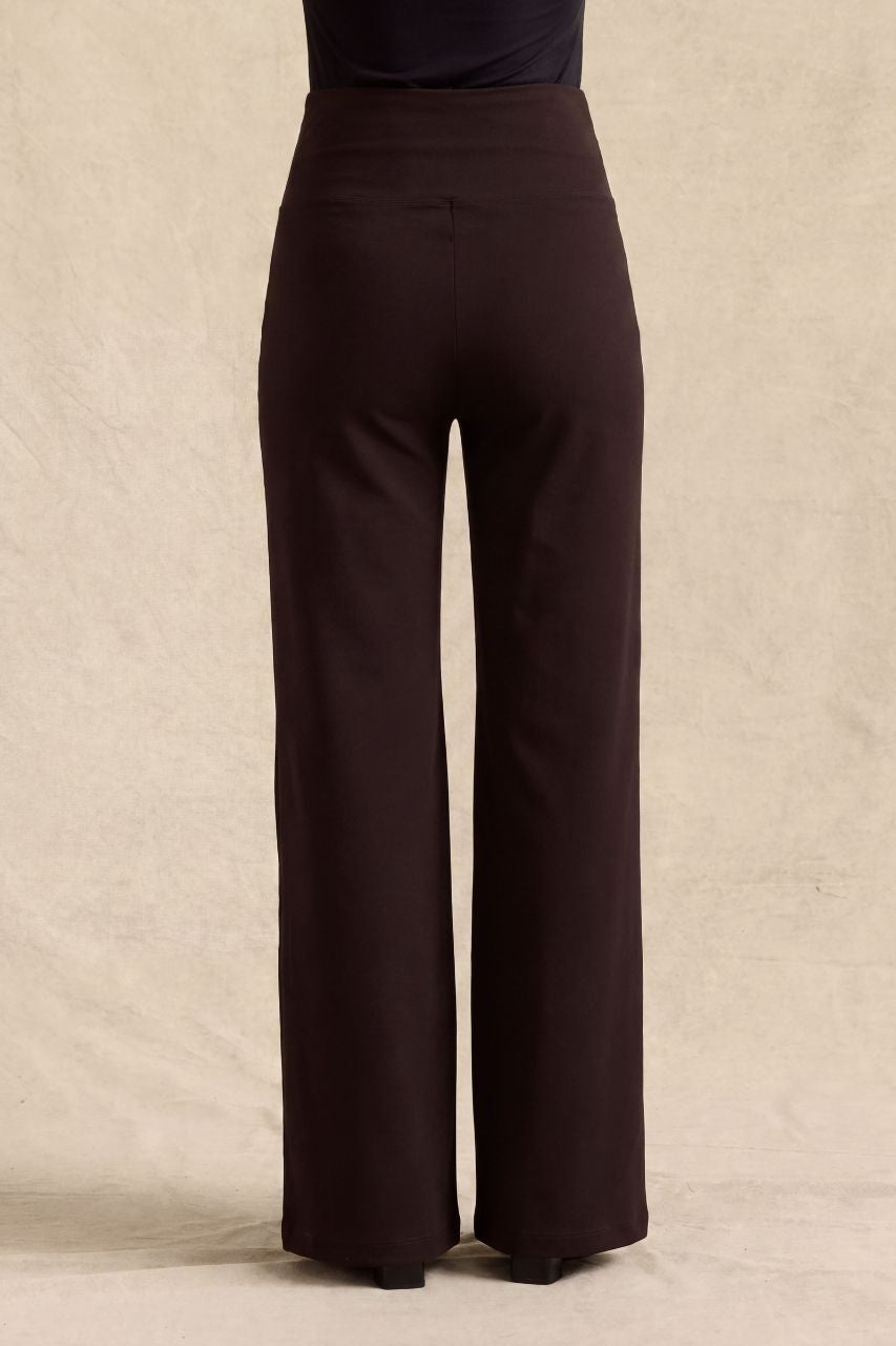 A pair of dark brown wide-leg trousers shown from the back on a model, against a textured beige backdrop.