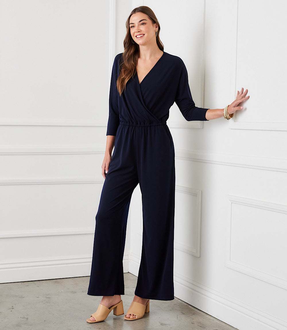 A woman modeling a navy blue wrap-style jumpsuit with three-quarter sleeves and wide legs, standing against a white paneled wall.