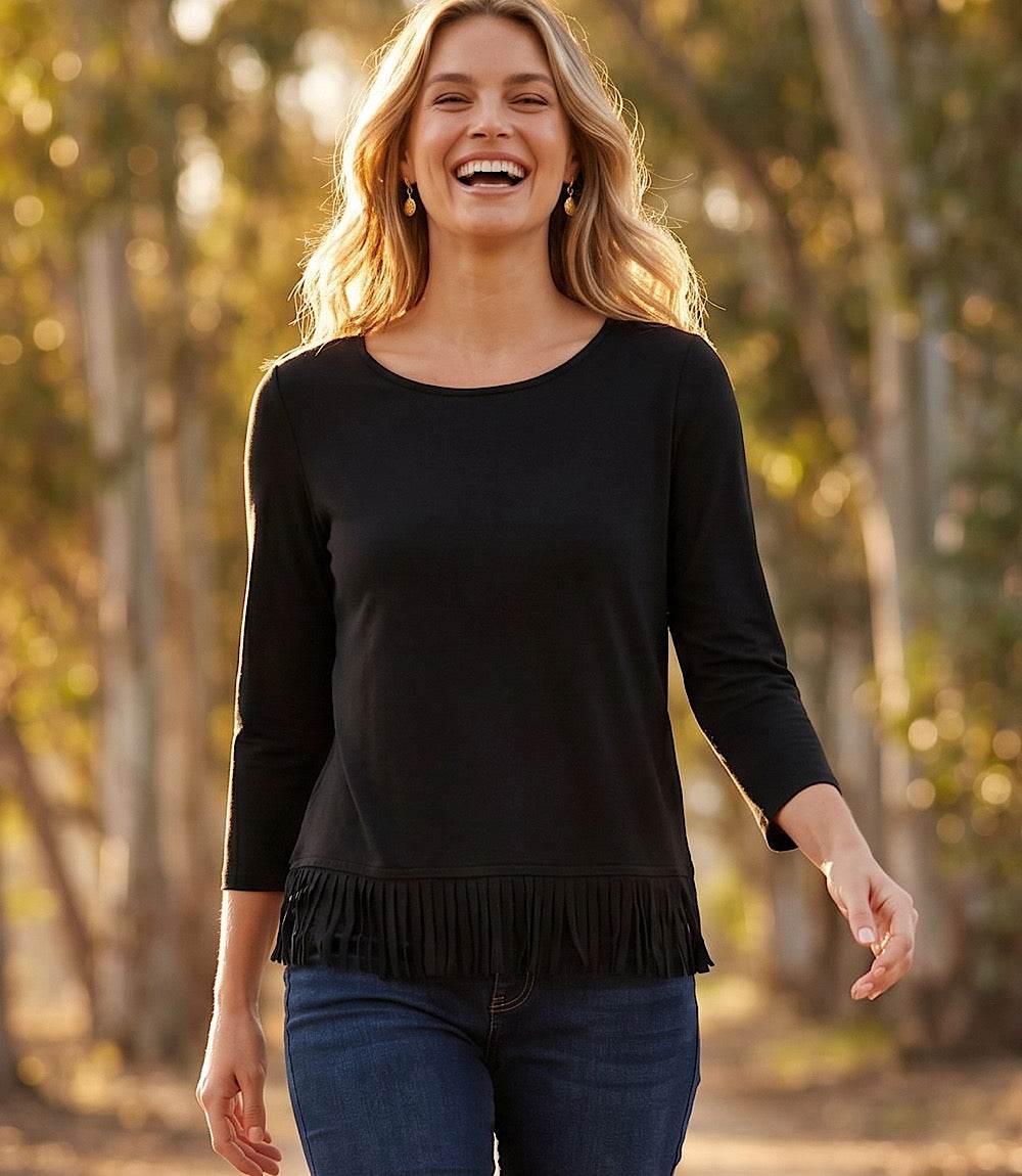 A woman smiling outdoors wearing a black long-sleeve top with a fringe hem and dark blue jeans, set against a blurred background of trees.