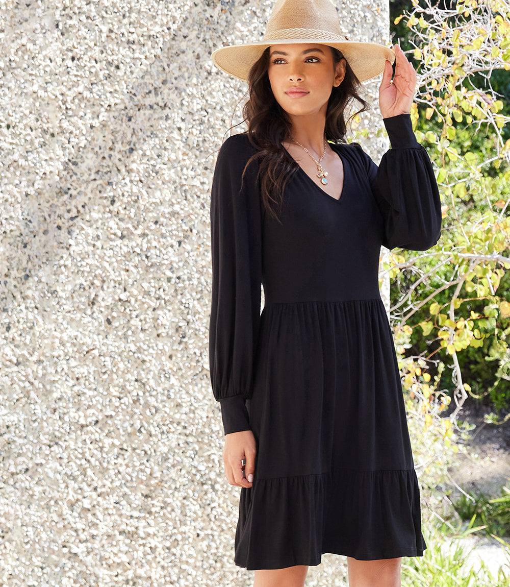 A woman wearing a black long-sleeve tiered mini dress and a beige wide-brim straw hat, standing outdoors against a textured light gray wall with green foliage on the right.