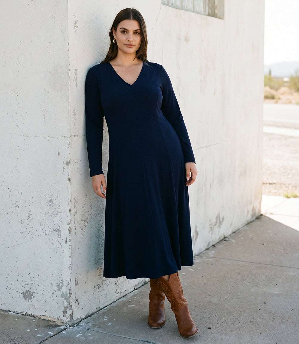 A woman wearing a long-sleeve navy blue midi dress and brown knee-high boots stands against a weathered white concrete wall.