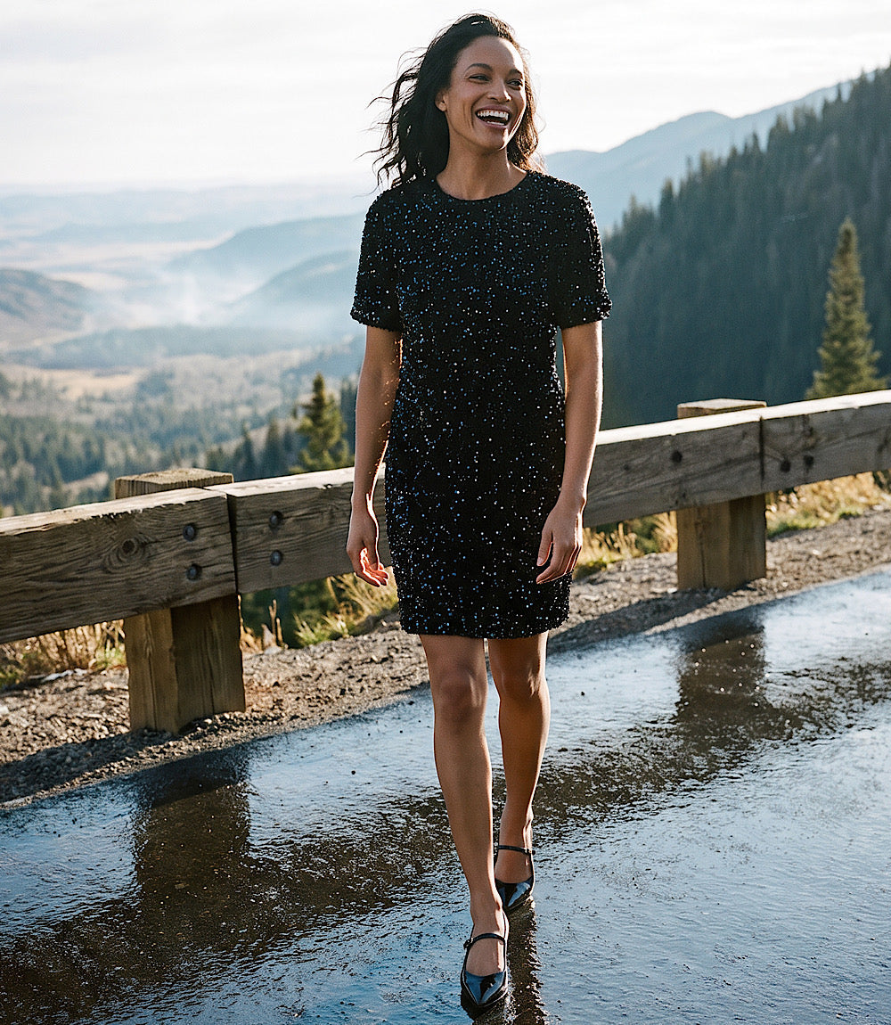 A woman wearing a short-sleeve black sequined dress walks along a wet road with a wooden guardrail and a mountain landscape in the background.