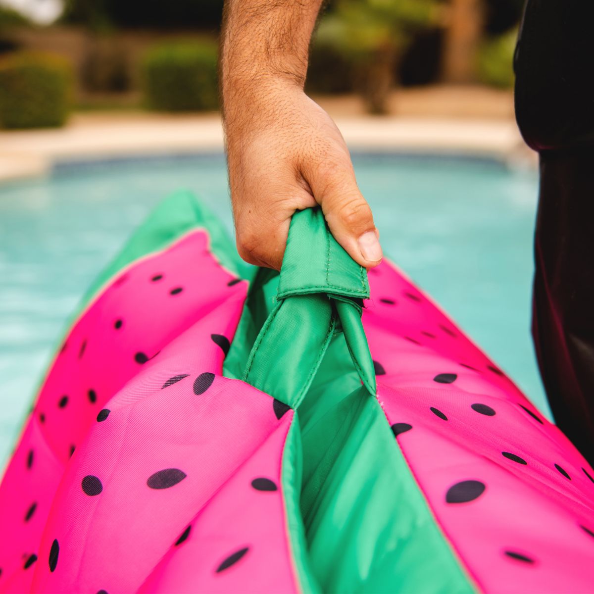 Hand holding a green inflatable pool toy shaped like a watermelon by a pool.