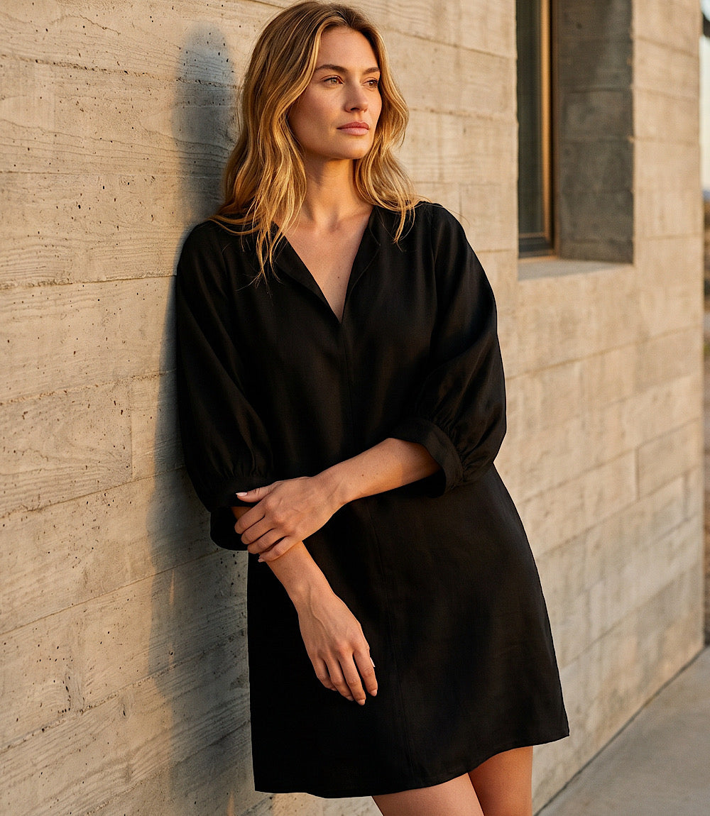 A woman wearing a black long-sleeve mini dress leans against a textured concrete wall outdoors.