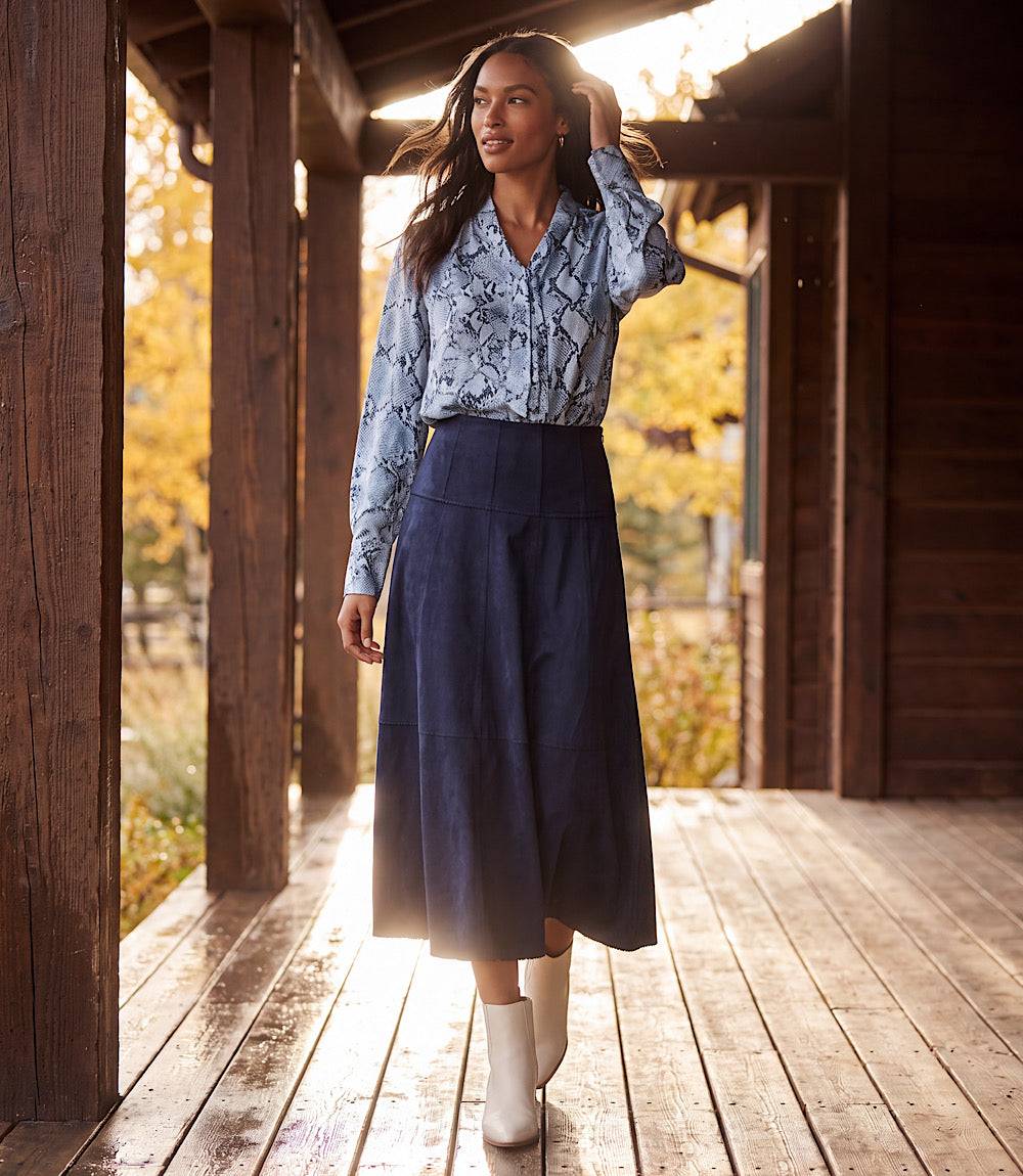 A woman stands on a wooden porch wearing a light blue snakeskin print long-sleeve shirt and a dark navy midi skirt, paired with white ankle boots. The background features warm autumn foliage visible through the porch beams.