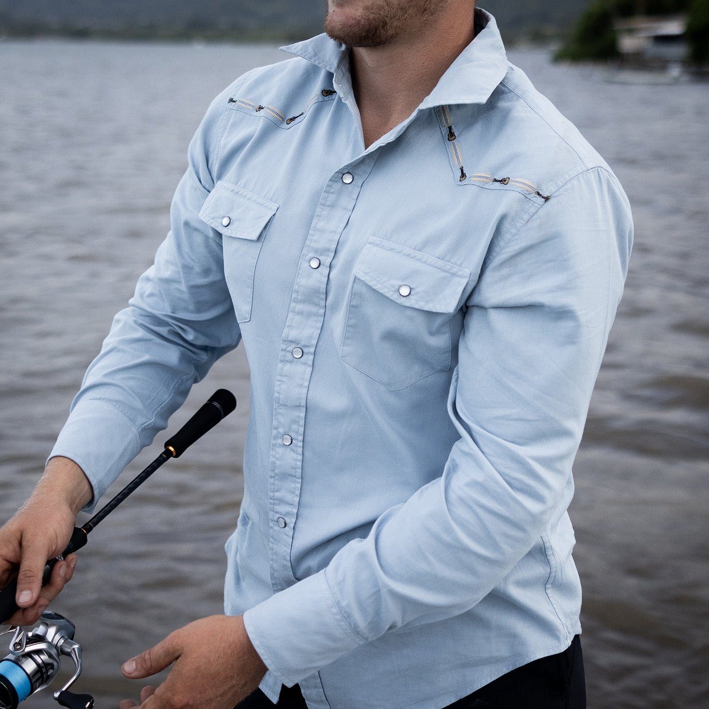 Man wearing a light blue denim shirt by a body of water, holding a fishing rod.