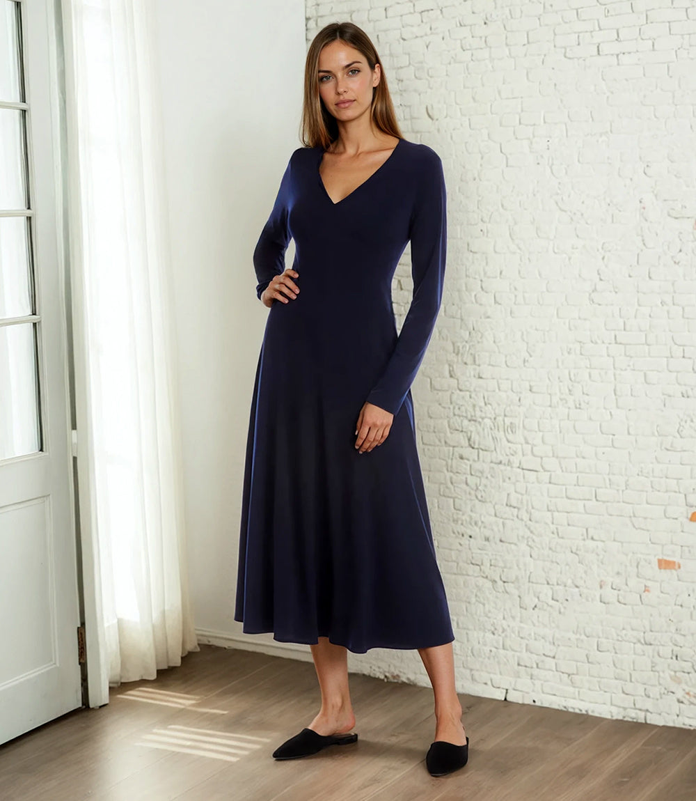 A woman modeling a long-sleeve navy blue midi dress with a V-neckline, standing in a bright room with a white brick wall and light wood floor.