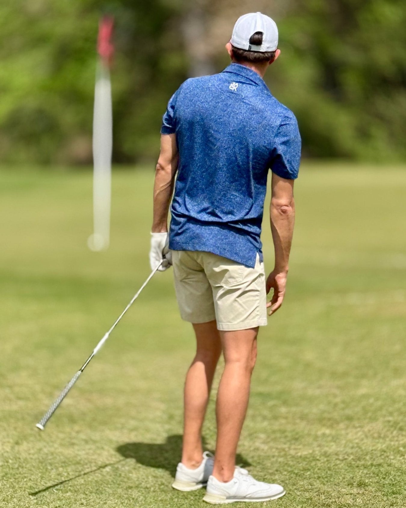 A man on a golf course seen from behind, wearing a blue patterned short-sleeve polo shirt, beige shorts, a white cap, and white golf shoes, holding a golf club.