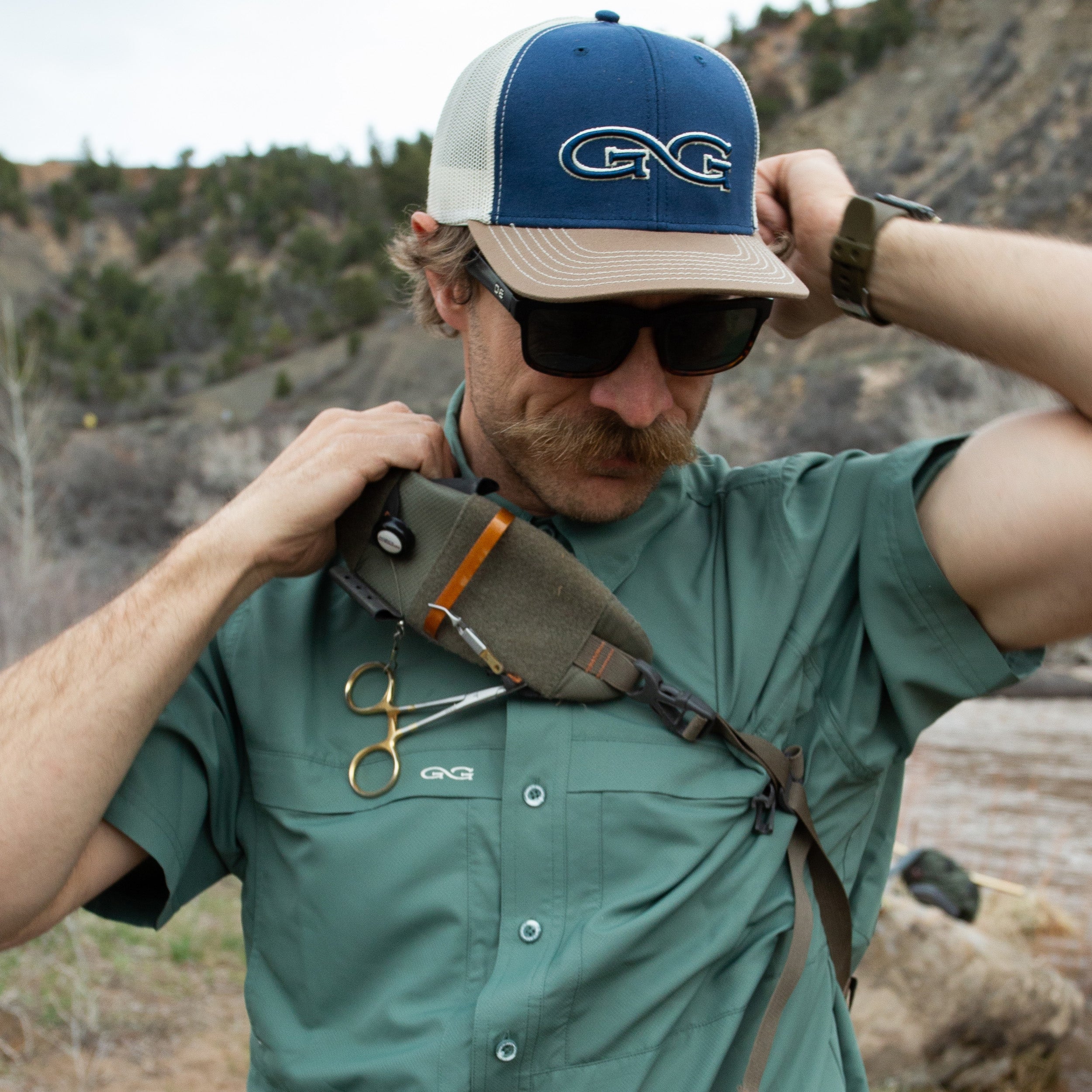 Man outdoors wearing a cap and sunglasses, with a backpack and tools on his chest.