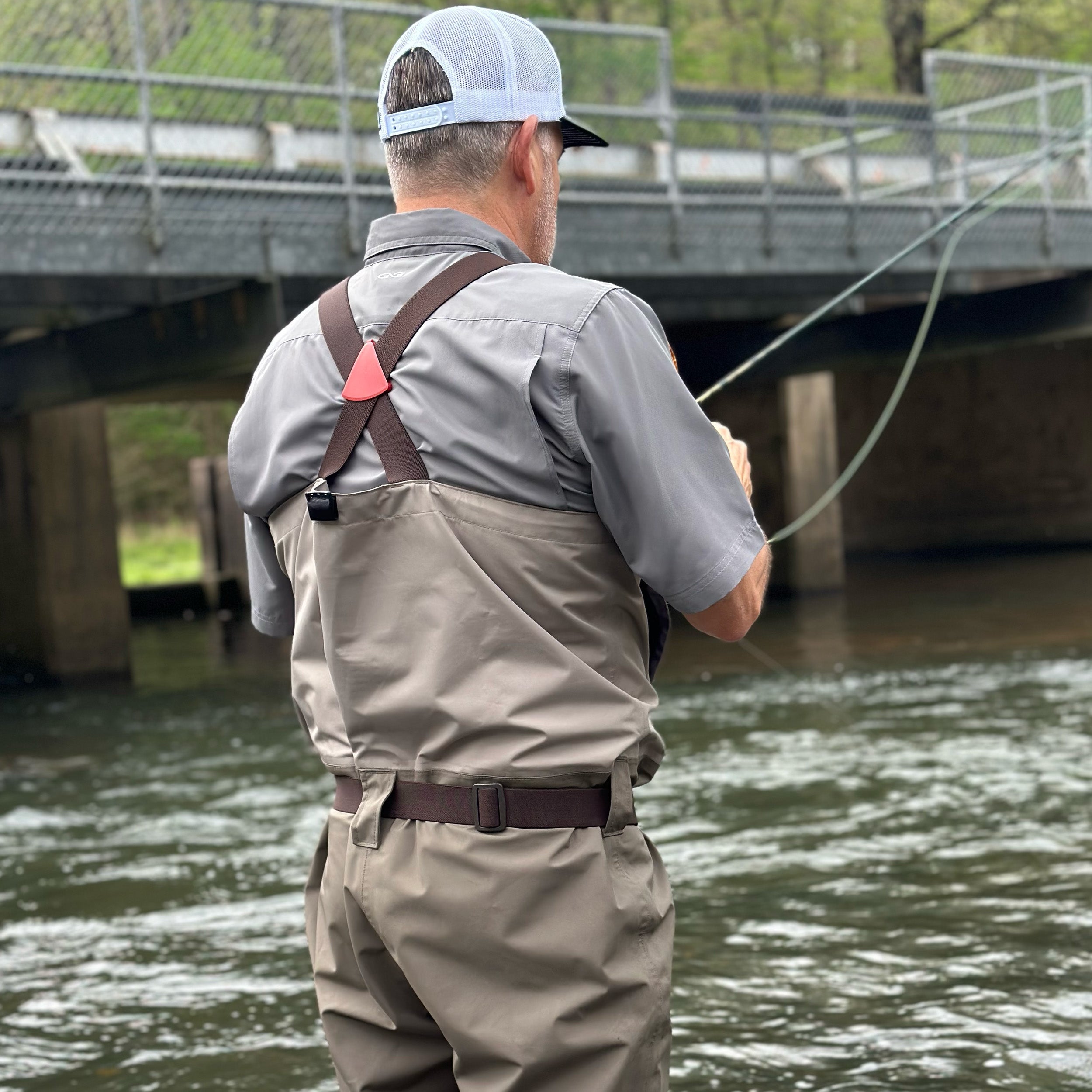 Man in fishing attire standing by a river with a bridge in the background