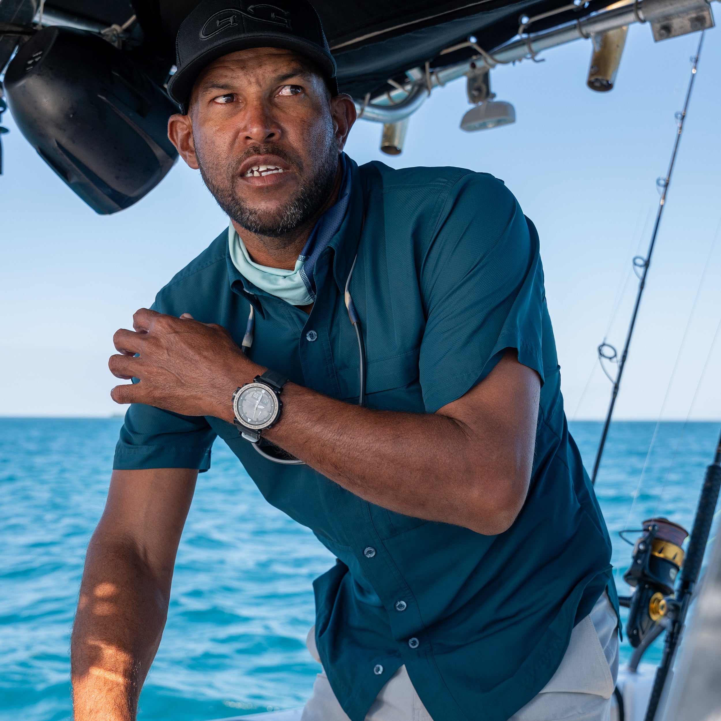 Man on a boat with fishing equipment, wearing a teal shirt and black cap, with blue water and sky in the background.