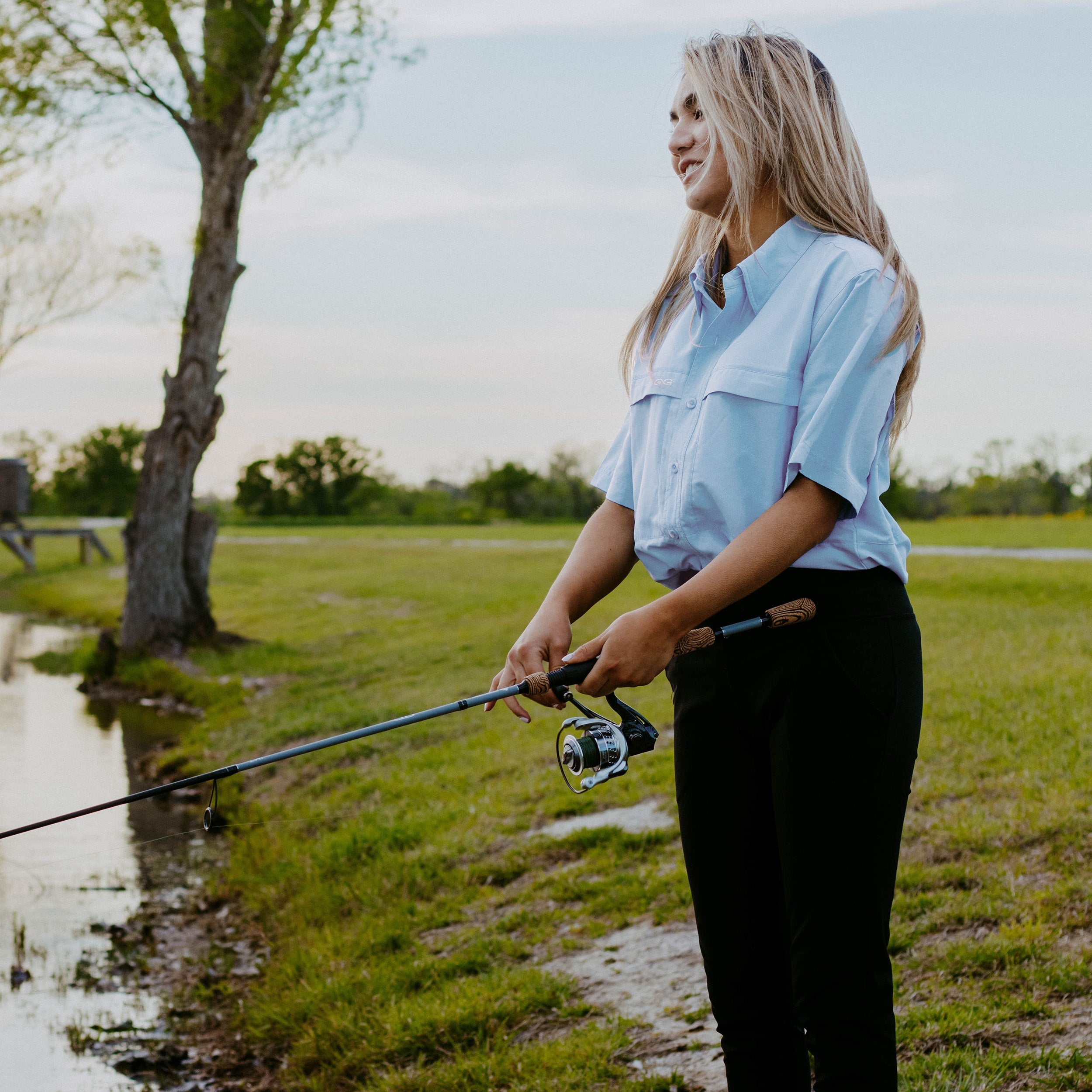 Woman holding a fishing rod by a pond with trees and grass in the background