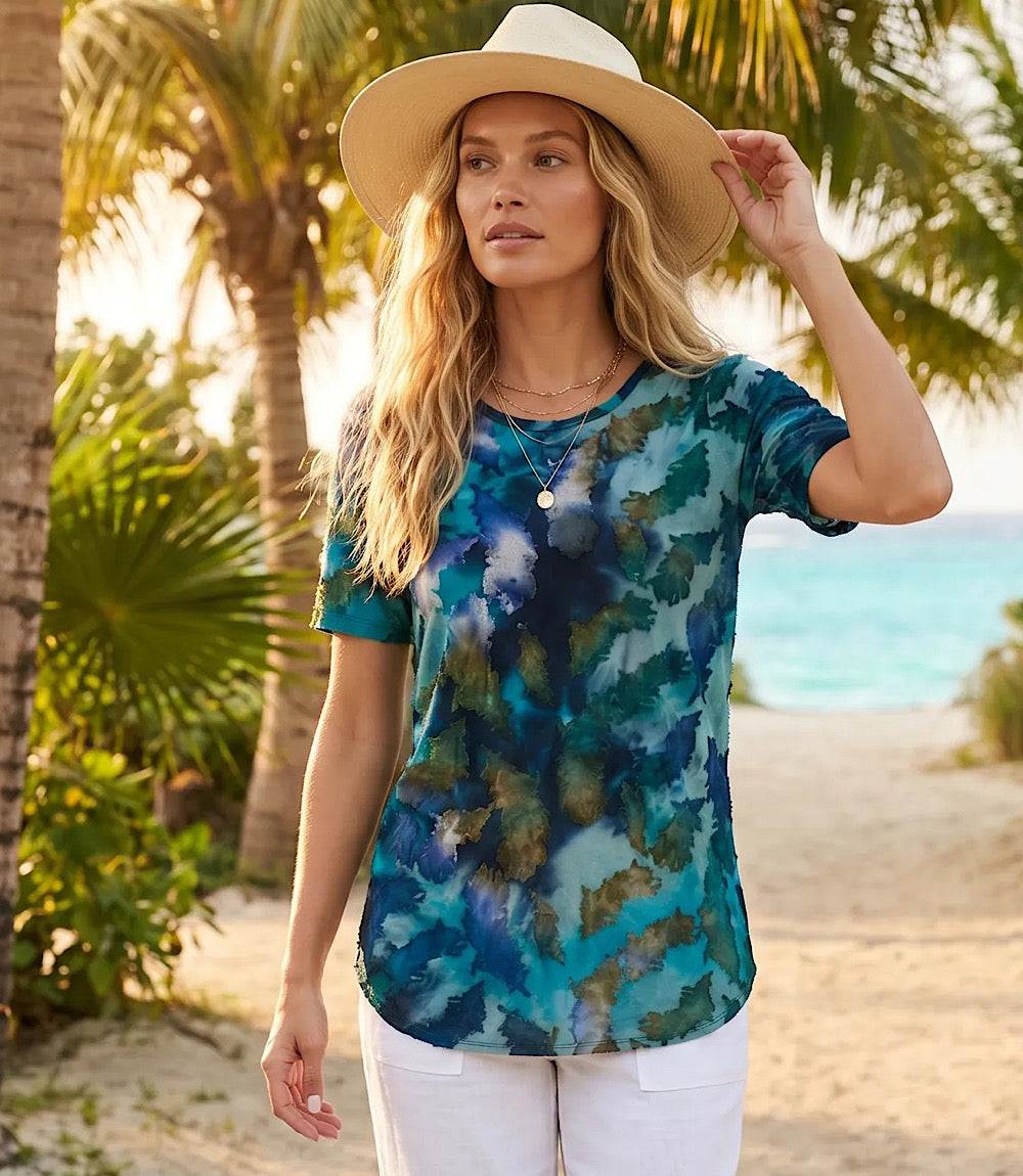 Woman wearing a colorful tie-dye shirt and wide-brimmed hat on a beach.
