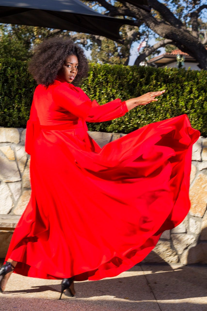 A woman outdoors wearing a flowing bright red maxi dress with long sleeves and black heels, captured mid-motion as the dress swirls around her against a hedge and stone wall background.