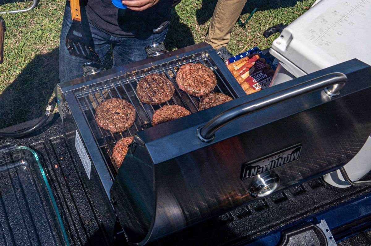 Hamburgers cooking on a portable grill attached to a vehicle, with a cooler in the background.
