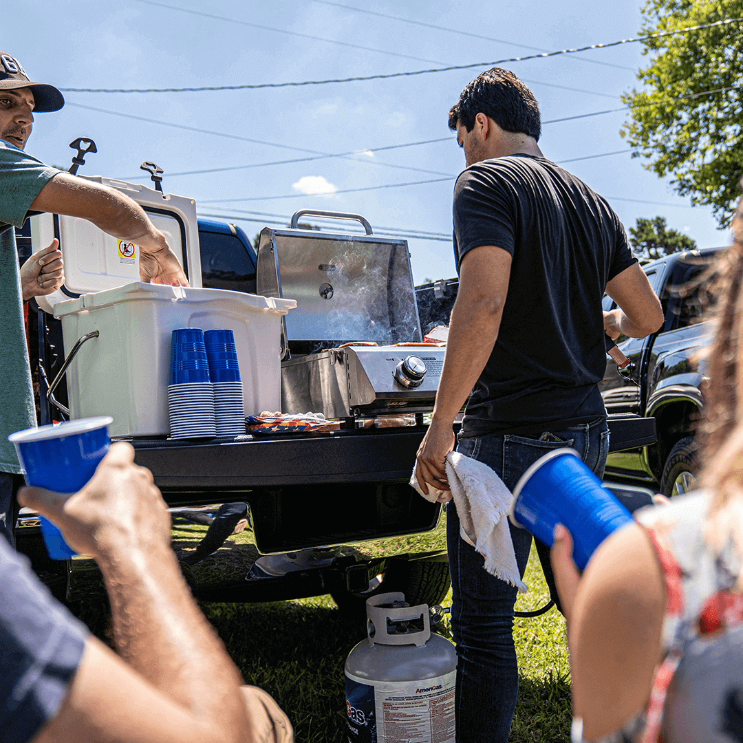 People around a grill with blue cups and a cooler in an outdoor setting