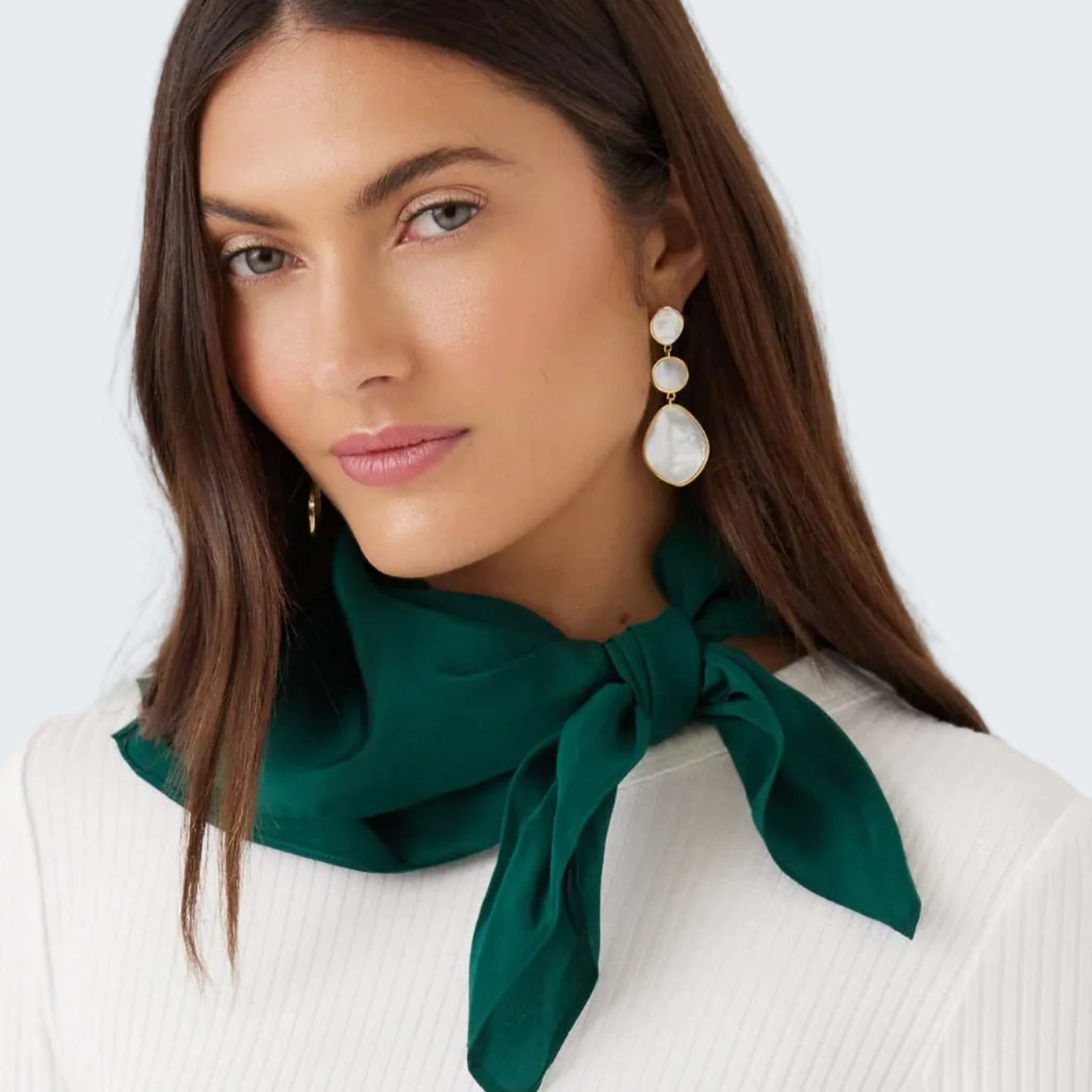 A woman wearing a dark green silk scarf tied in a knot at the collar, paired with white pearl drop earrings and a white ribbed top, set against a plain light gray background.