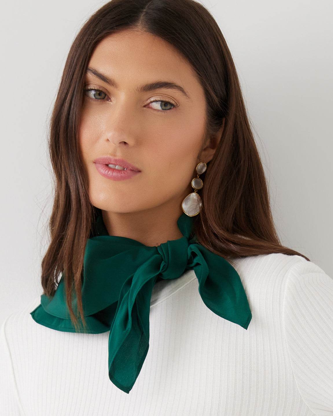 A woman with long brown hair models a dark green satin neck scarf tied in a large bow at the collar, paired with a white ribbed top and dangling pearl earrings against a light gray background.