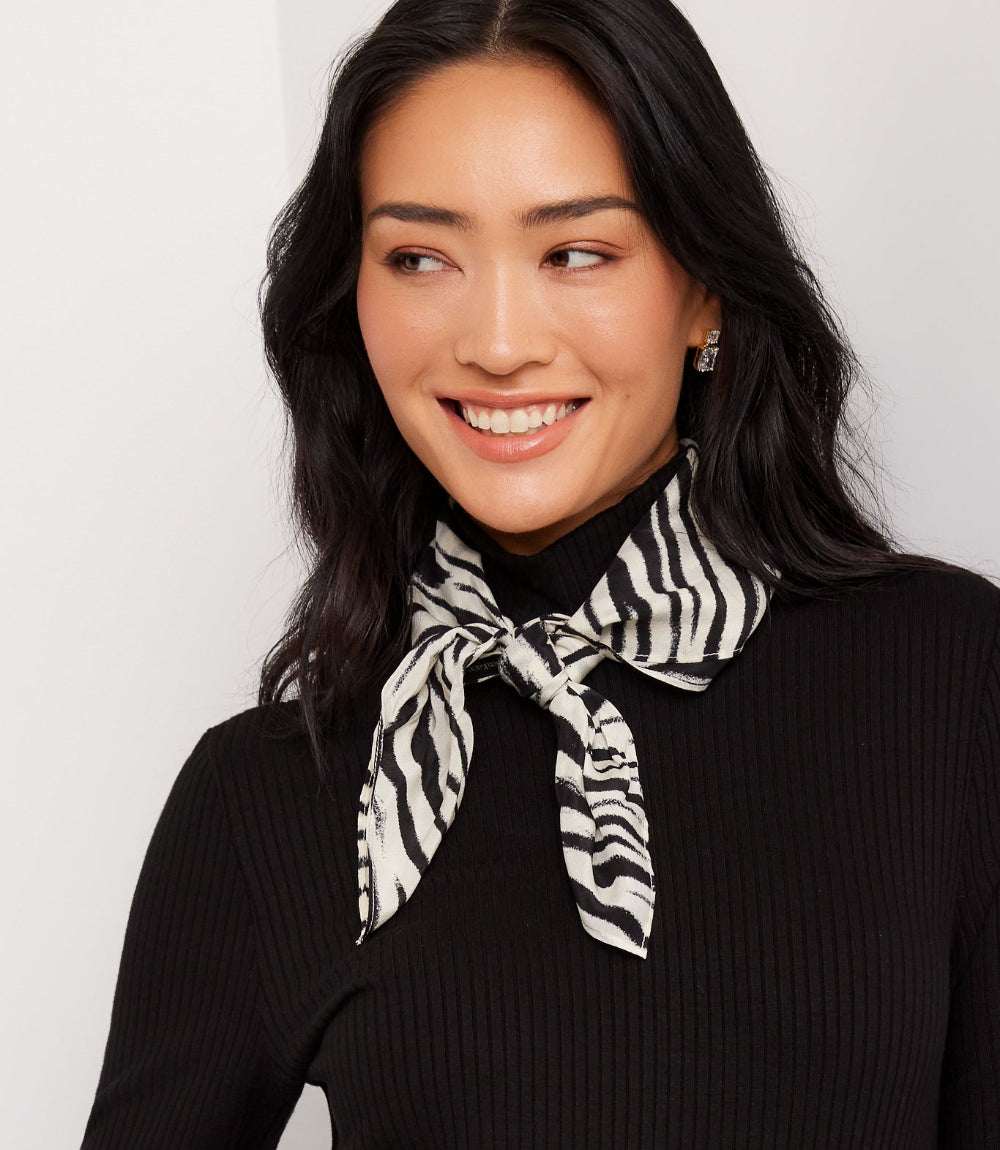 A woman with long black hair wearing a black ribbed turtleneck sweater and a black and white zebra-print neck scarf tied at the collar, shown against a plain light gray background.