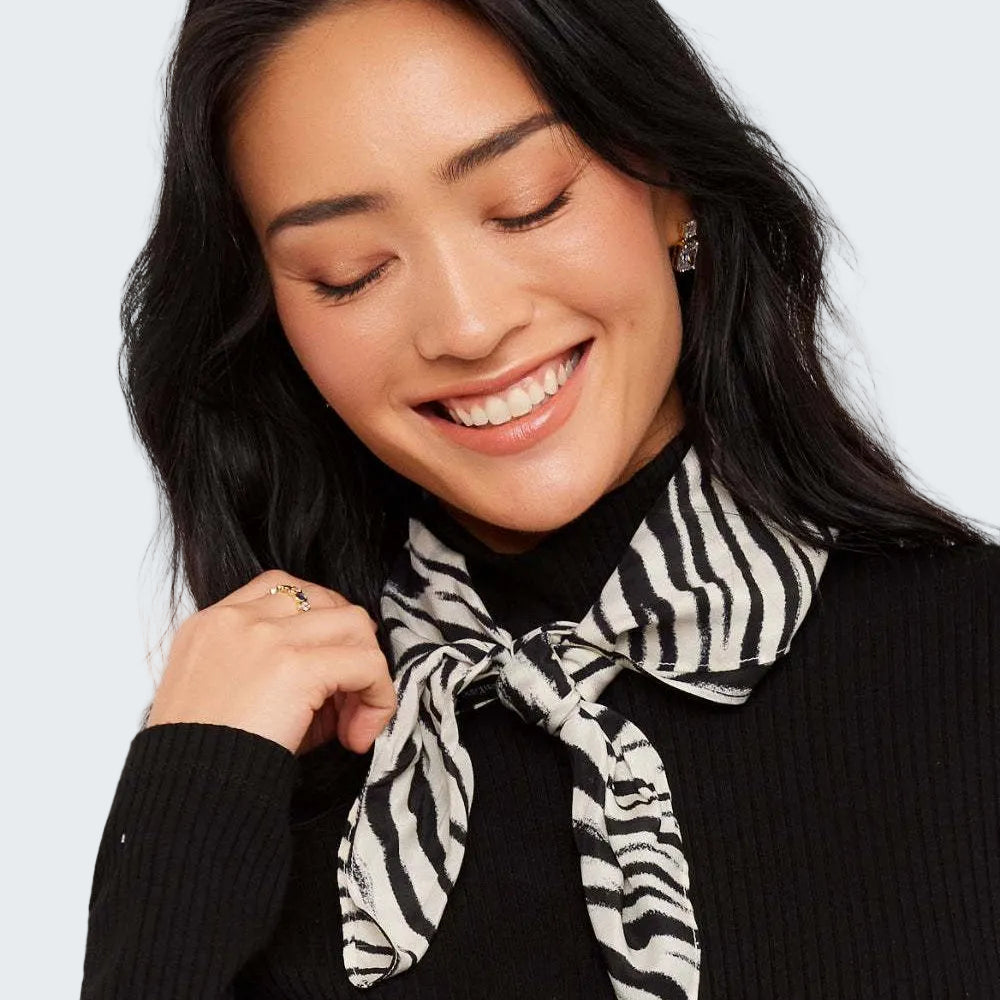 A woman with long black hair smiling and looking down, wearing a black ribbed turtleneck sweater and a black and white zebra-print neck scarf tied at the collar.