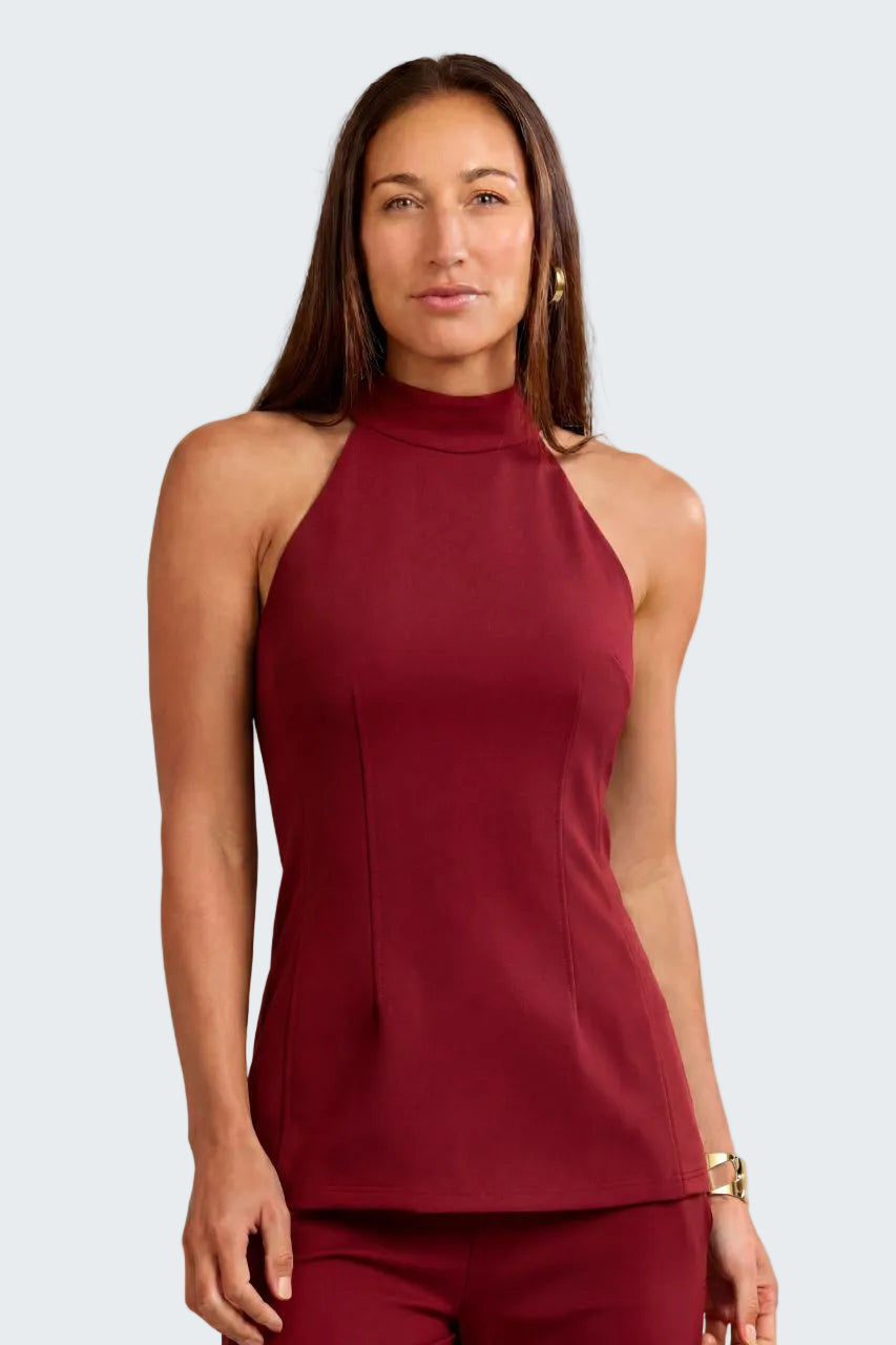 A woman modeling a sleeveless high-neck top and matching pants in a deep burgundy color, set against a neutral textured beige background.