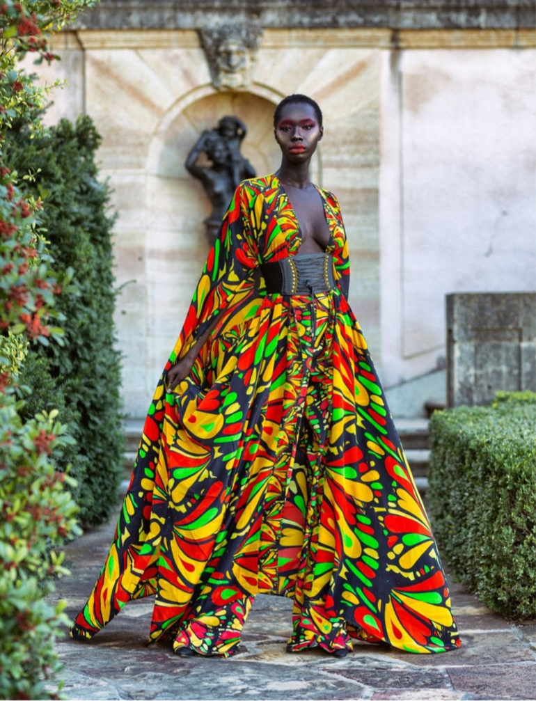 Person wearing a colorful patterned dress standing in an outdoor setting with a statue and greenery.