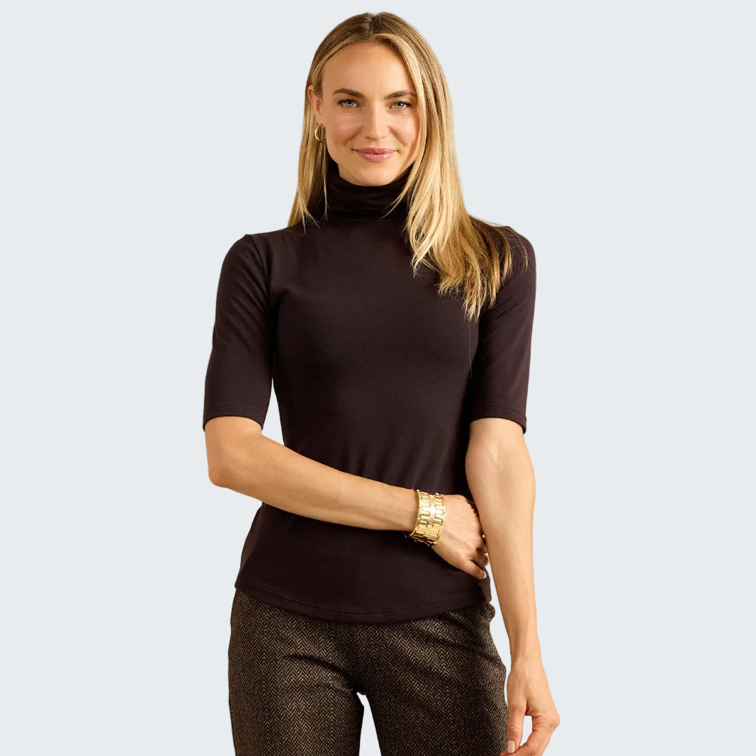 A woman modeling a short-sleeve, high-neck mock neck top in a very dark plum or eggplant color, paired with textured dark pants, against a neutral beige textured wall.