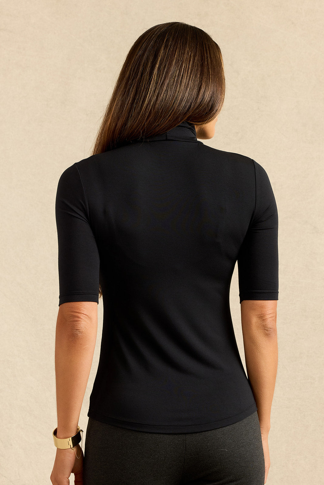 A woman viewed from the back wearing a fitted black short-sleeve athletic top with a high mock neck, set against a plain beige textured background.