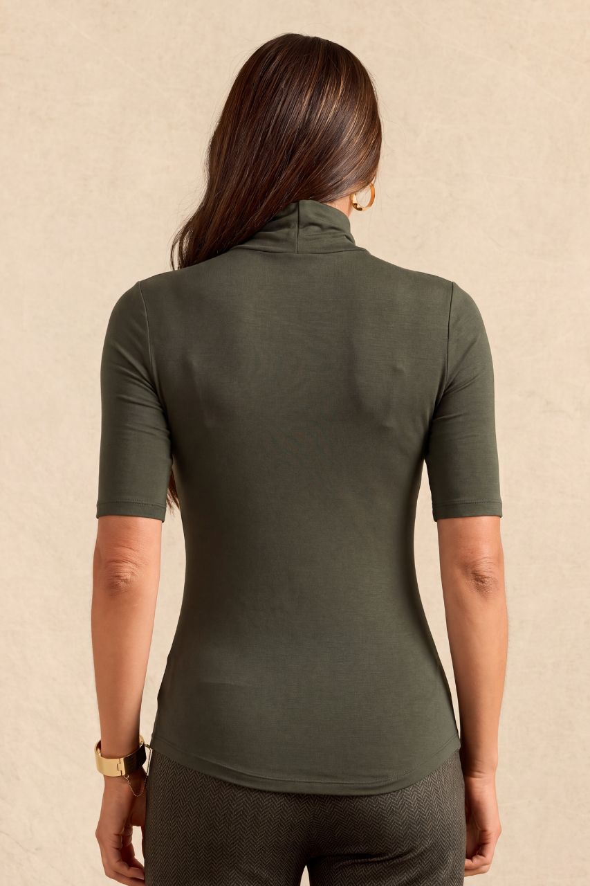 A woman viewed from the back wearing a fitted short-sleeve turtleneck top in olive green, paired with dark gray pants, against a textured beige background.