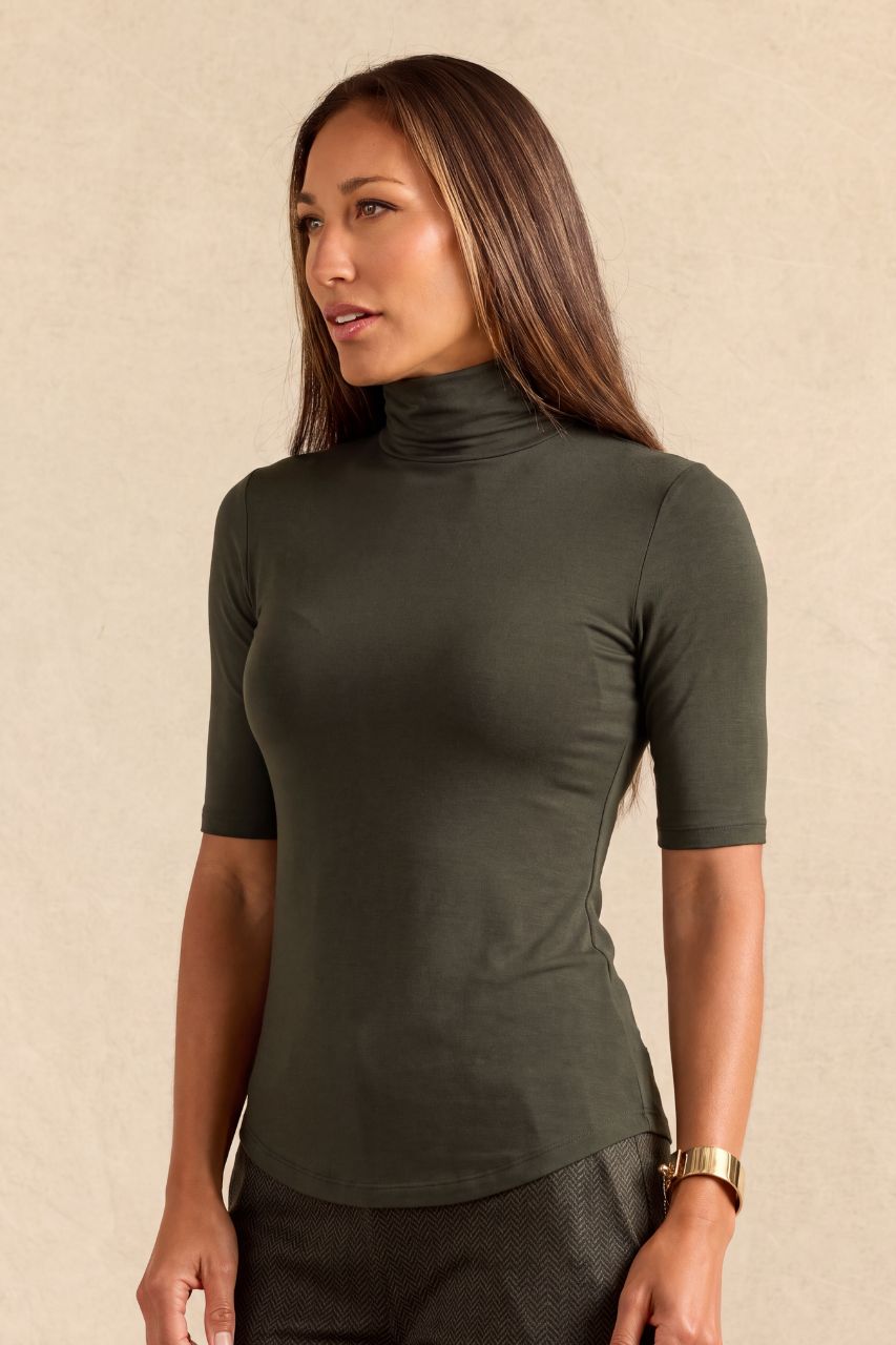 A woman wearing a fitted short-sleeve turtleneck top in dark olive green, standing against a plain beige textured wall.