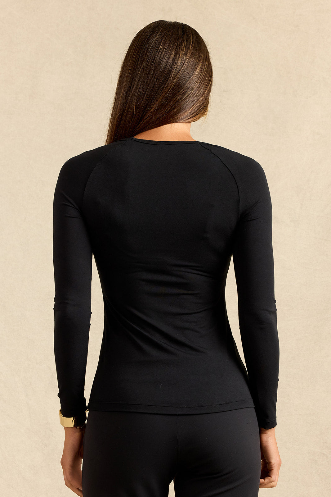 A woman viewed from the back wearing a fitted black long-sleeve athletic top and black leggings against a warm beige textured background.