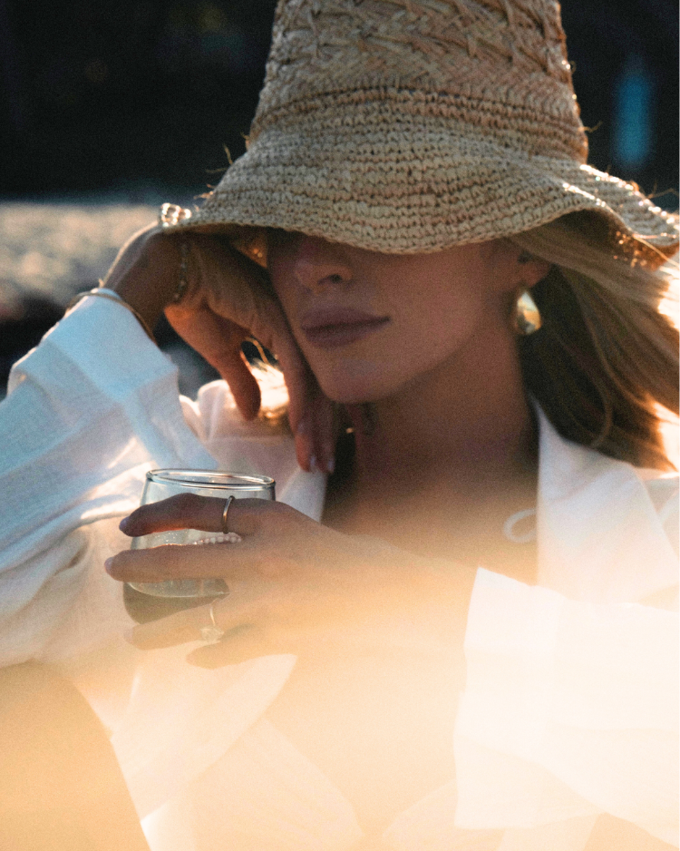 Woman wearing a straw hat and white top, holding a glass outdoors.