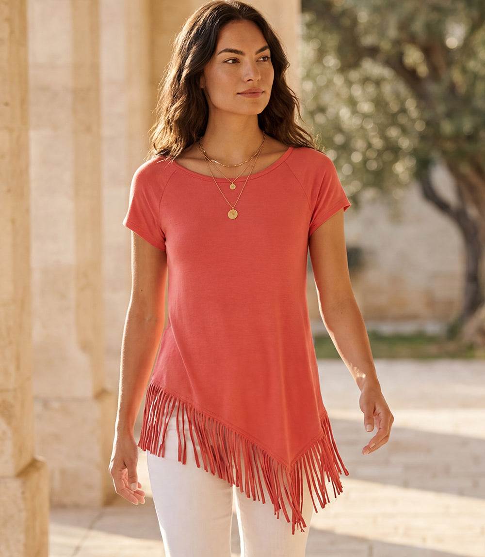 A woman wearing a coral pink short-sleeve tunic top with fringe trim at the hem, paired with white pants, standing outdoors in front of a sunlit stone building.