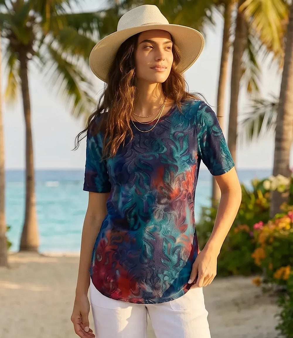 A woman wearing a short-sleeve t-shirt with a vibrant teal, blue, and red abstract floral pattern, styled with a wide-brimmed straw hat and white pants, standing on a tropical beach with palm trees and the ocean in the background.