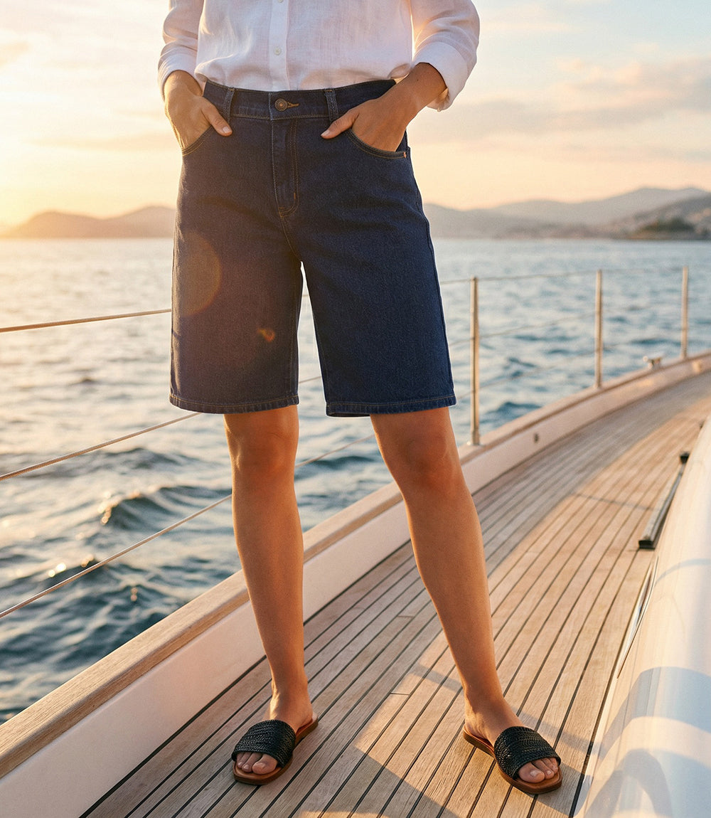 A person standing on a yacht deck by the ocean at sunset, wearing dark blue denim shorts, a white button-up shirt, and black slide sandals.