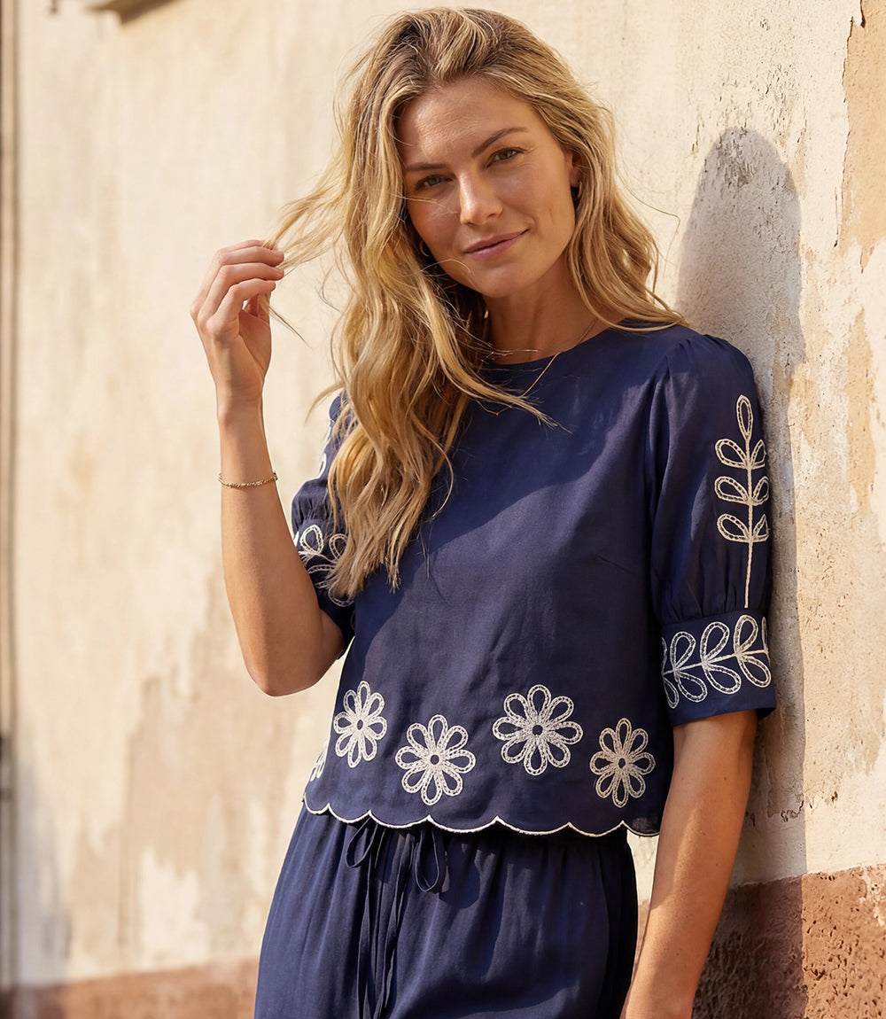 Woman wearing a navy blue top with white floral embroidery against a stone wall.