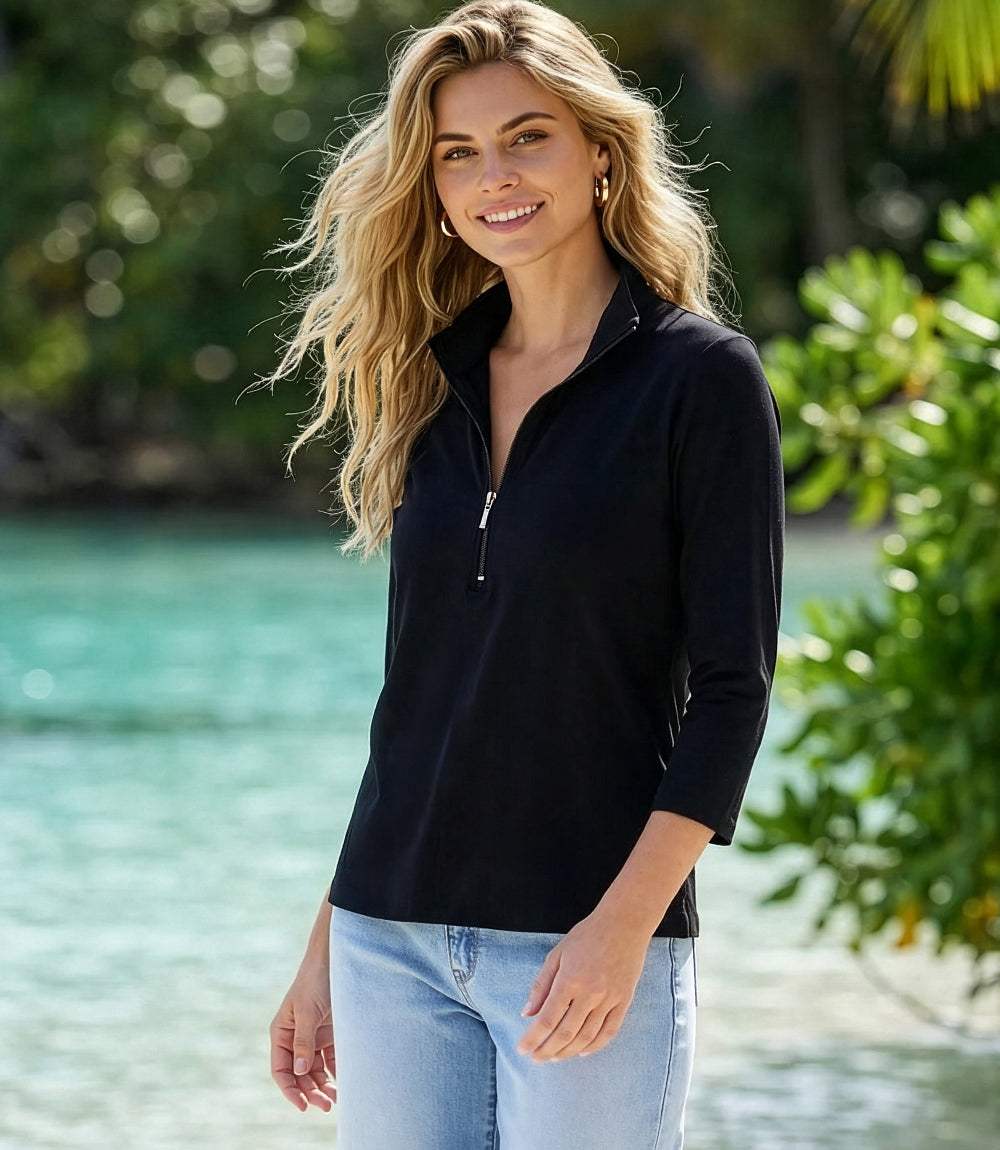 A woman with blonde hair stands outdoors by the water wearing a black three-quarter zip long-sleeve top and light blue jeans.