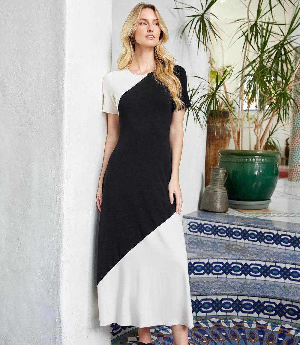 A woman modeling a short-sleeve color-block midi dress with black, white, and beige sections, standing in a Mediterranean-style interior next to a tiled table and potted plants.