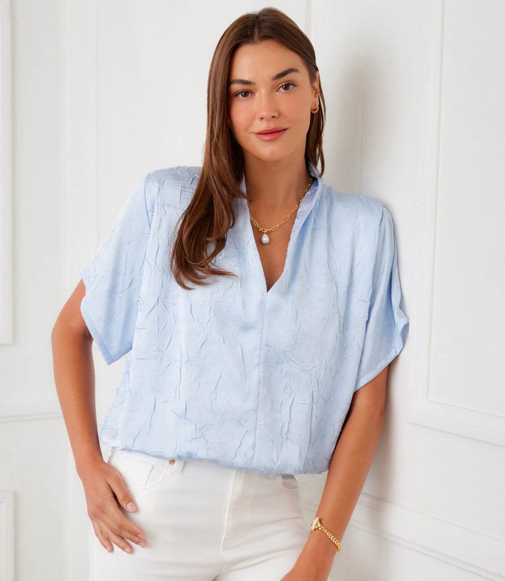 A woman wearing a light blue textured short-sleeve blouse and white trousers, standing against a white paneled wall.