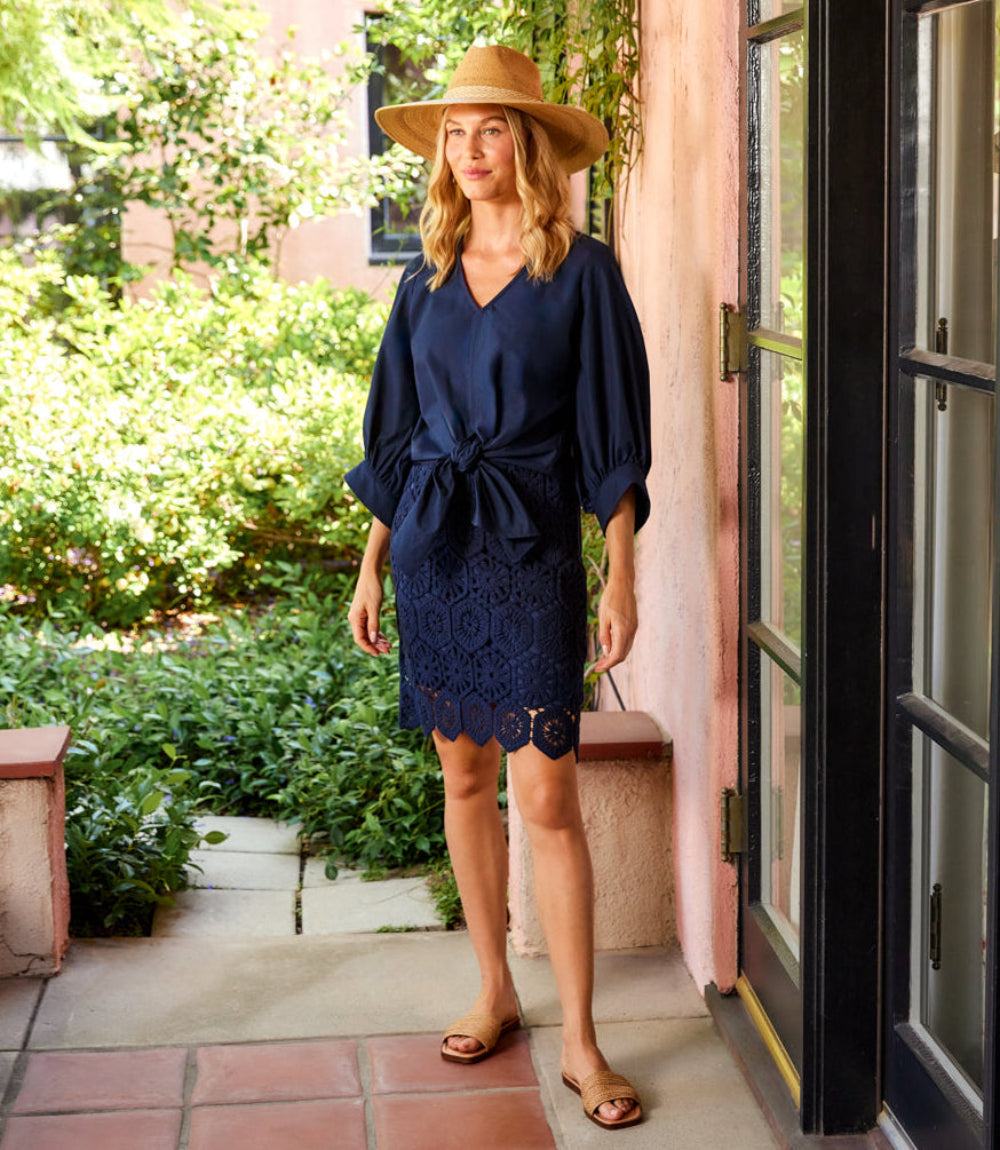 A woman wearing a navy blue lace and tie-front blouse with a matching lace mini skirt and a wide-brim straw hat, standing on a tiled porch with a pink stucco wall and green garden in the background.