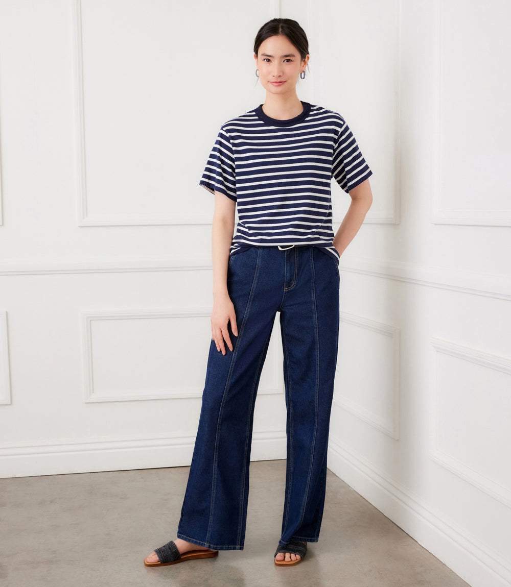 Woman wearing a striped shirt and blue pants standing against a white paneled wall.