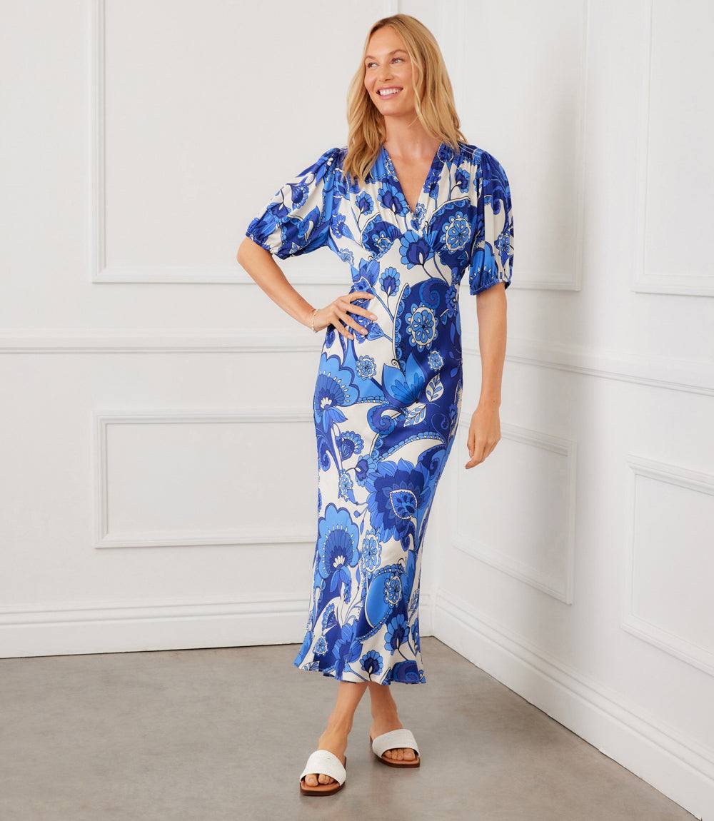 Woman wearing a blue floral dress standing against a white paneled wall.