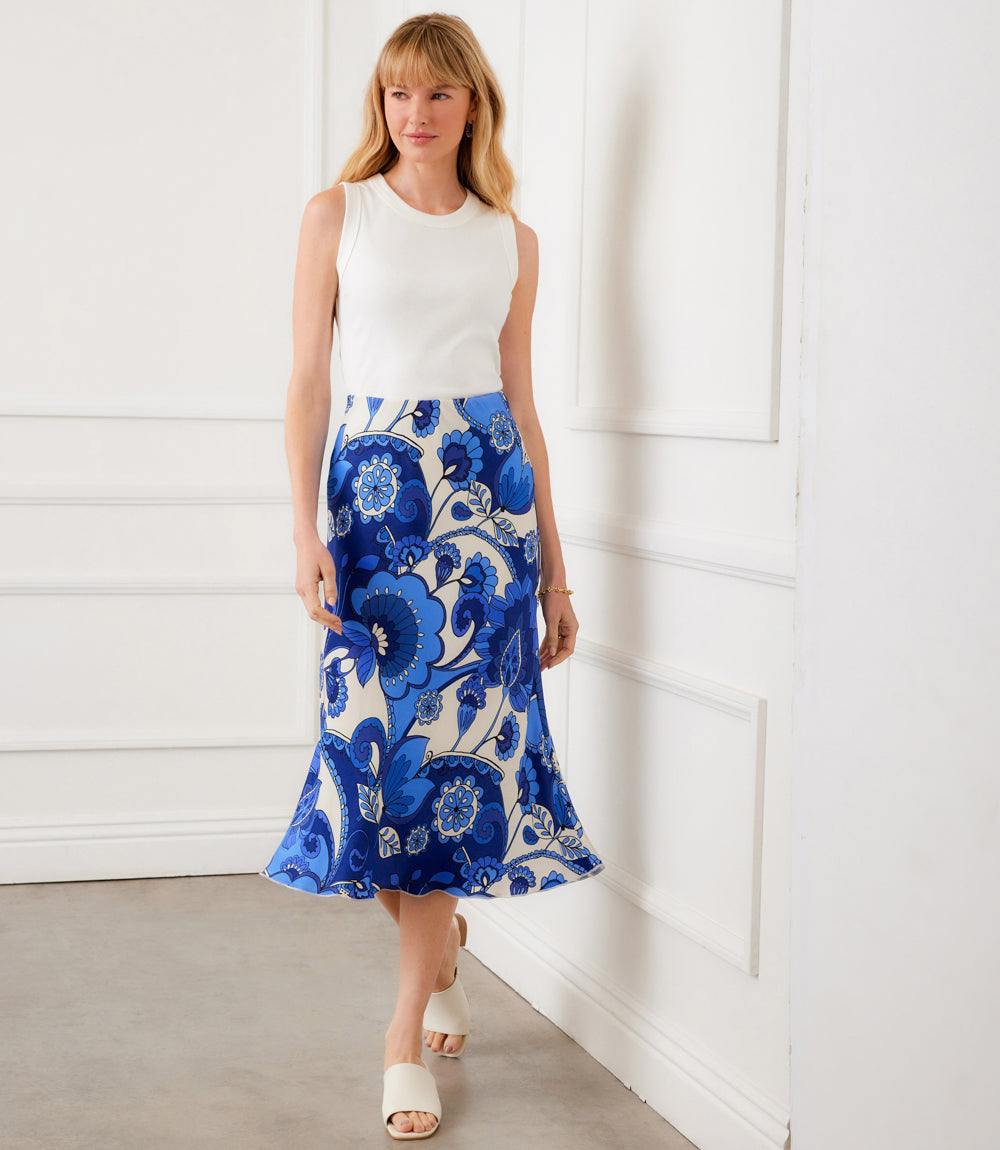 Woman wearing a white sleeveless top and blue floral skirt against a white wall.