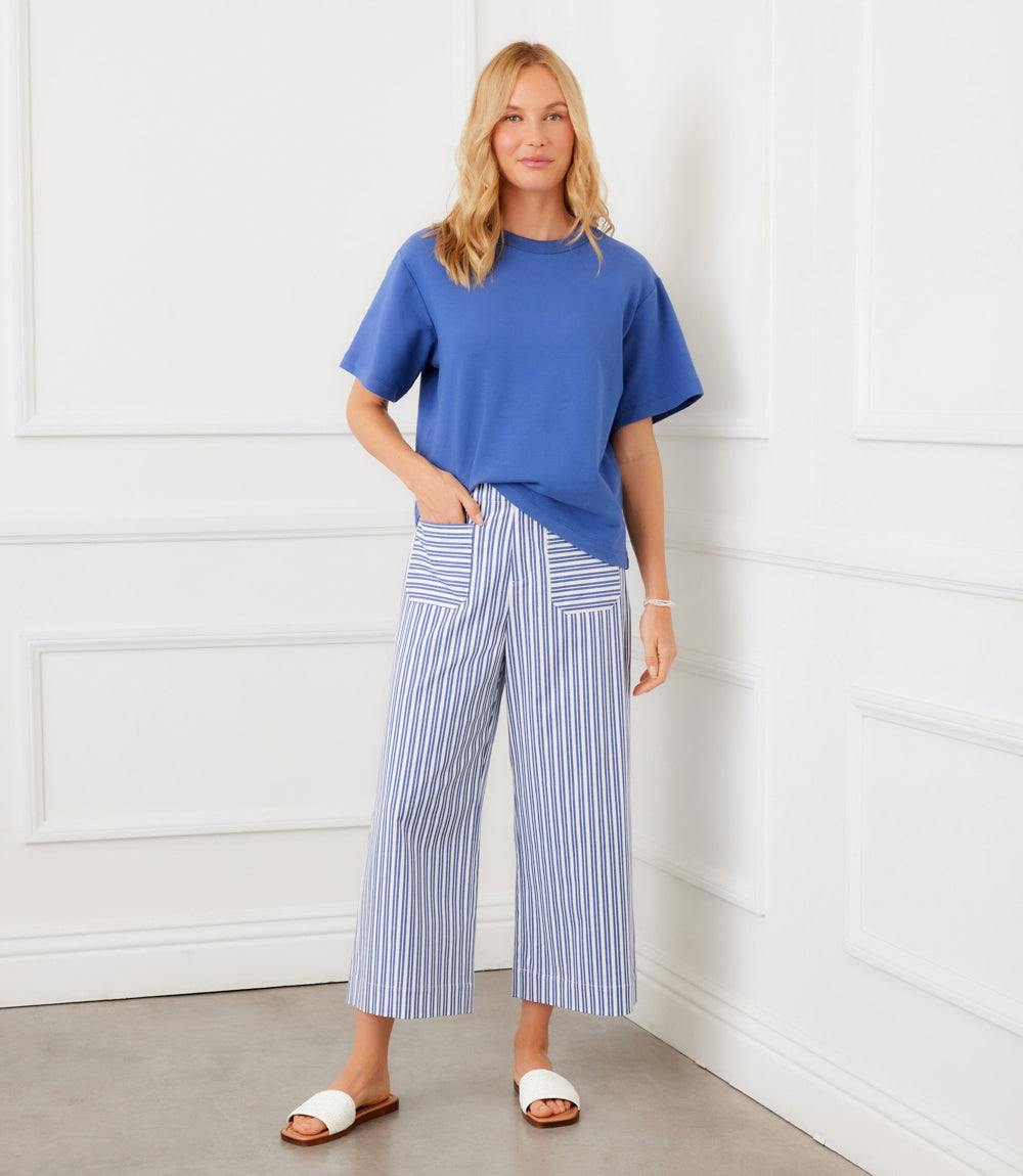 Woman wearing a blue top and striped pants against a white paneled wall.