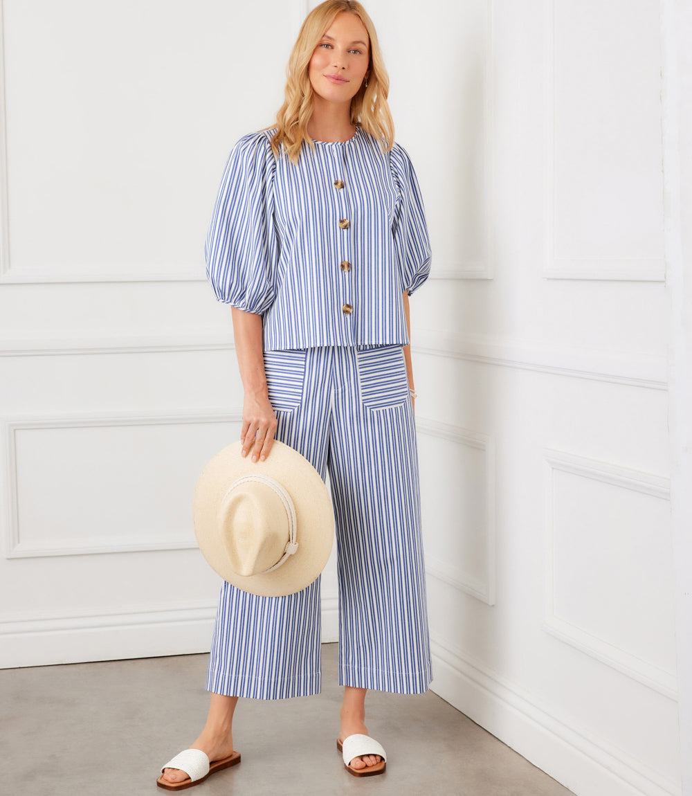 Woman wearing a blue and white striped outfit holding a beige hat against a white wall.