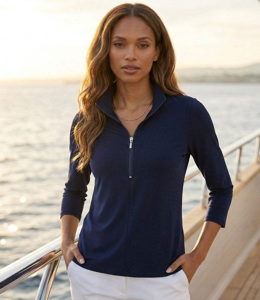 A woman wearing a navy blue quarter-zip long-sleeve top and white pants, standing on a yacht deck with water and a warm sunset sky in the background.