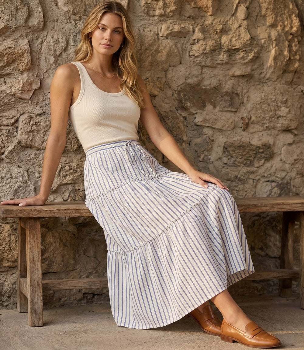 A woman sits on a rustic wooden bench against a stone wall, wearing a cream ribbed tank top and a tiered midi skirt with thin vertical blue stripes. She is wearing tan leather loafers.