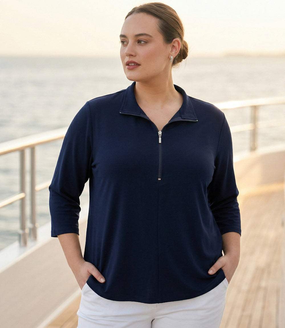 A woman wearing a navy blue quarter-zip long-sleeve top and white pants, standing on a wooden deck with the ocean in the background.